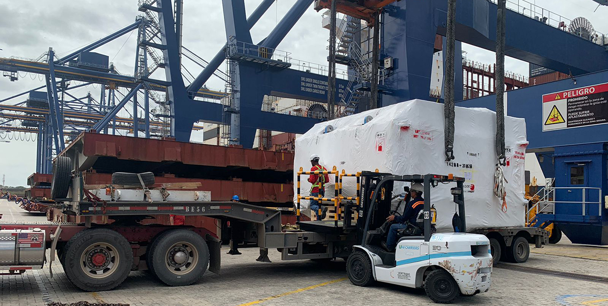 Trabajadores usando una carretilla elevadora para mover una carga cubierta en un muelle con grúas y contenedores al fondo.