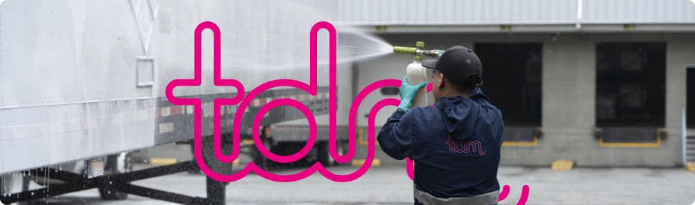 Una persona con ropa de trabajo limpia un vehículo con un rociador de alta presión; letras rosadas en primer plano.