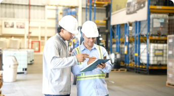 Dos trabajadores con cascos blancos revisan un formulario en un almacén.