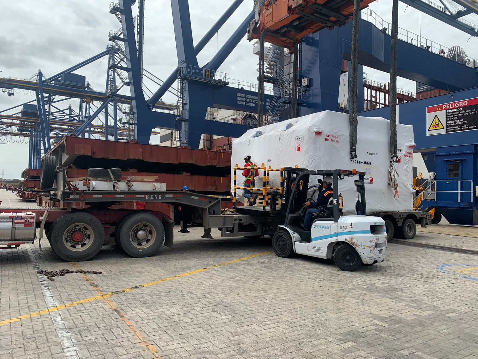Carretilla elevadora transportando una carga envuelta en plástico en un puerto, con grúas y camiones en el fondo.