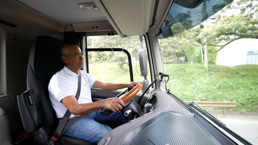 Hombre mayor conduciendo un camión, sostiene el volante y usa cinturón de seguridad; paisaje verde visible por la ventana.