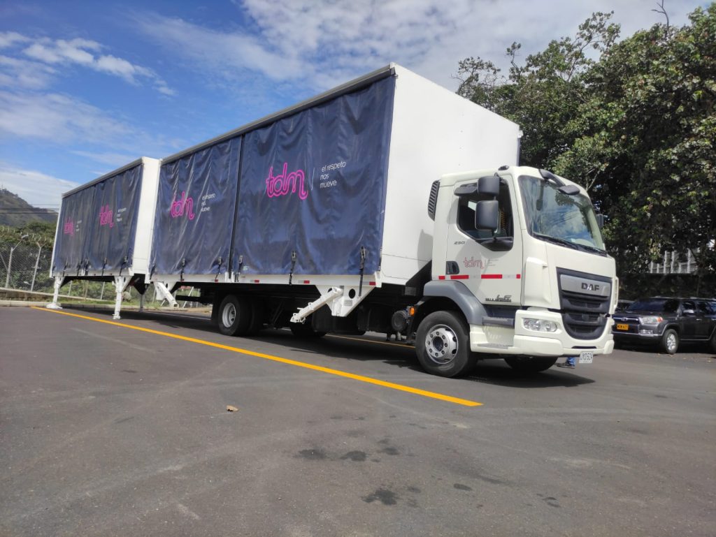 Camión blanco con remolque azul de lona, estacionado al aire libre en un día soleado.
