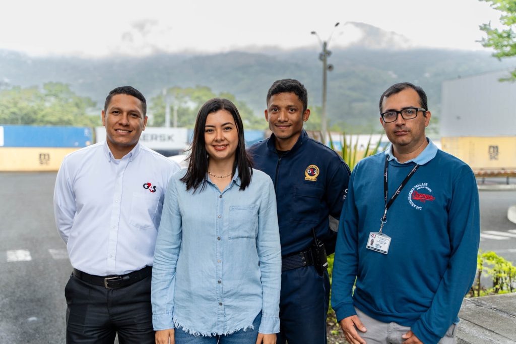 Cuatro personas de pie al aire libre, con montañas y contenedores de carga al fondo.