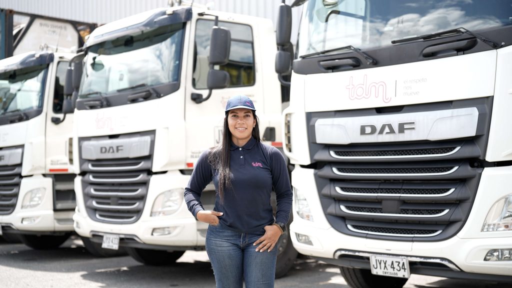 Una mujer con uniforme de trabajo sonríe frente a una fila de camiones DAF blancos estacionados.
